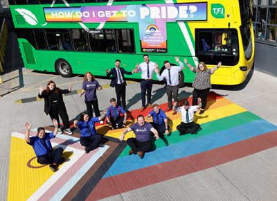 Image of Dublin Bus employees with hands raised and a green and yellow bus behind them, standing on Pride flag