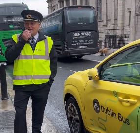 Image of chief inspector, Mark Drew on street talking on a handset with a Dublin Bus yellow and green car beside him